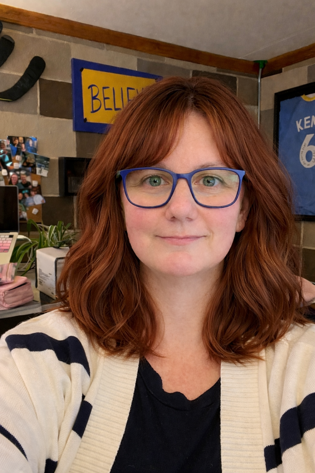Rebecca Lamont in her workspace with a 'BELIEVE' sign, hockey memorabilia, and plants behind her